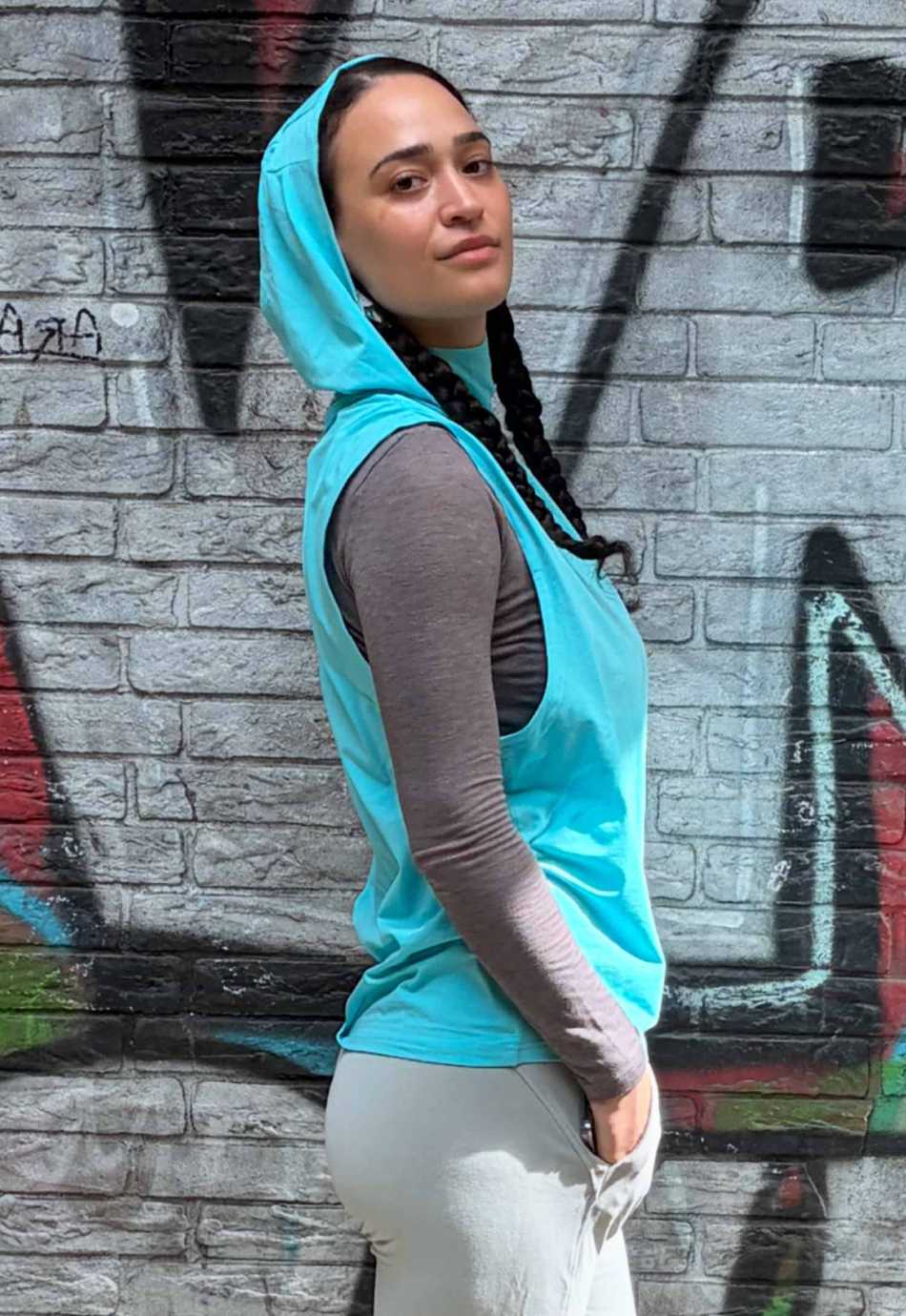 Kyle Hoodie ärmellos Unisex in Türkisblau – Seitenansicht vor einer Graffiti-Wand, Kapuze auf