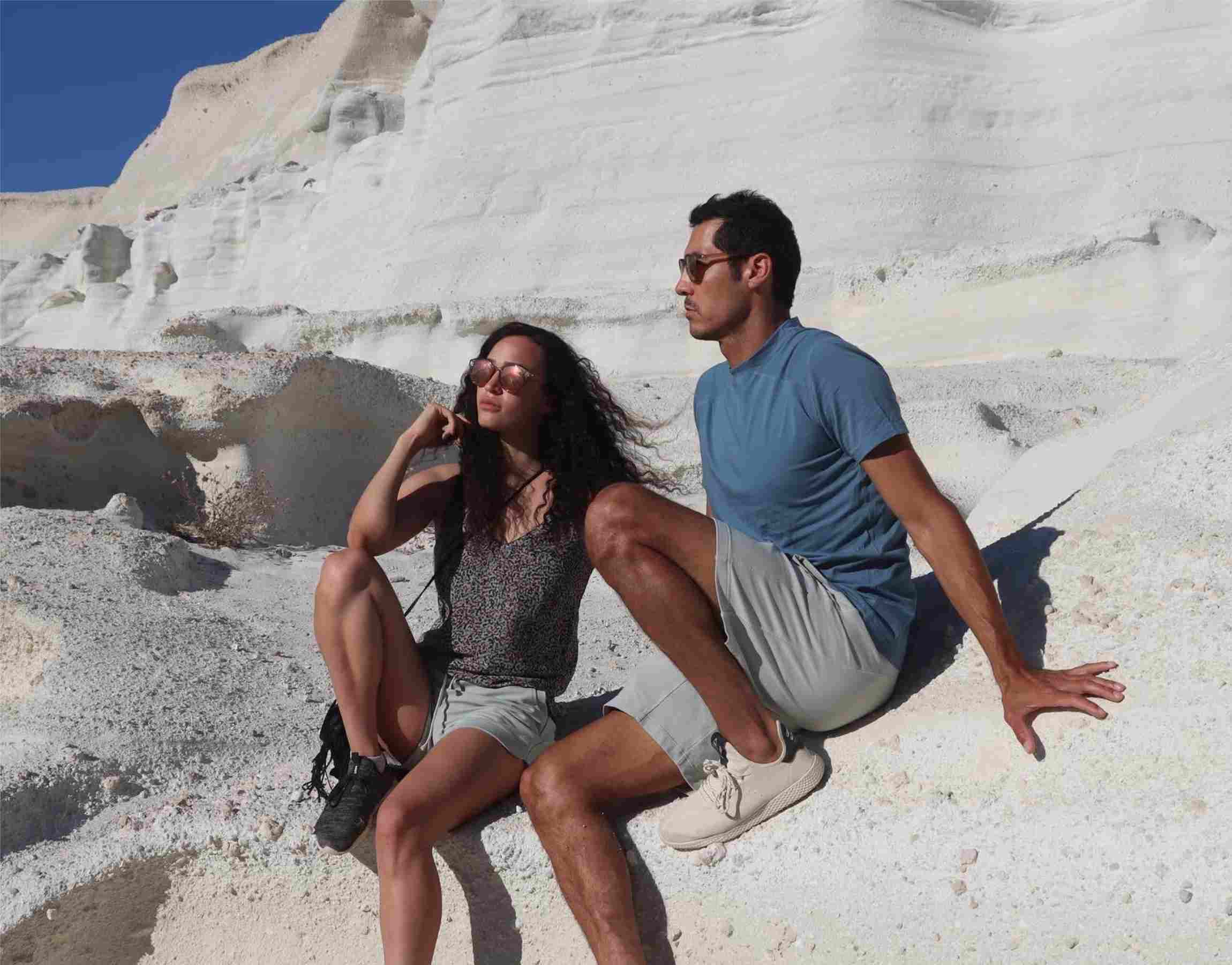 M23-Models in nachhaltiger Sportmode aus Bio-Baumwolle – Frau in umweltfreundlichen Activewear-Shorts und Mann im blauen Performance-T-Shirt – sitzen auf weißen Felsen in Milos, Griechenland.

